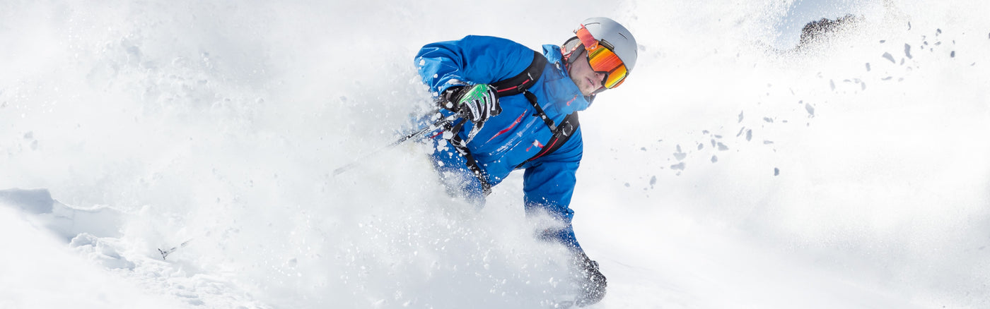 Man in a blue jacket, white helmet and orange snow goggles, skiing down a snow slope with a large snow cloud